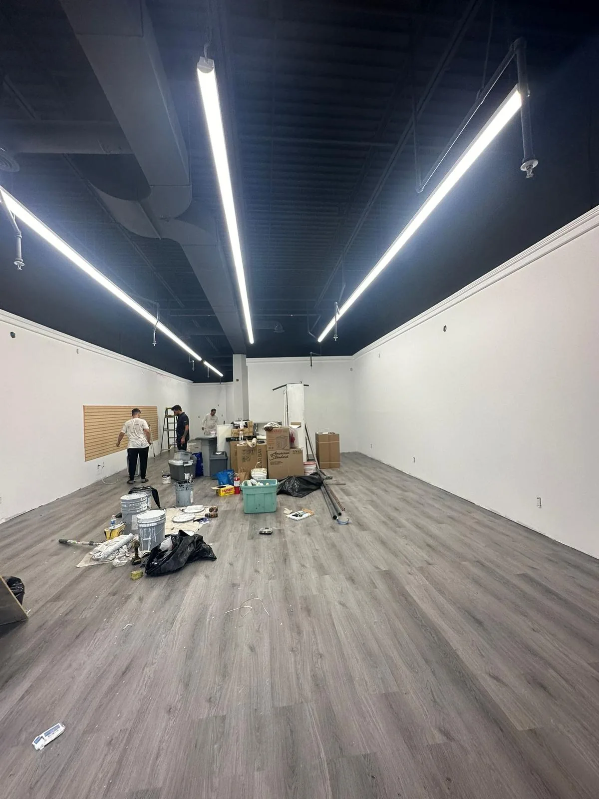 The same unit mid-project with new white walls, a ceiling sprayed black, linear lighting and new grey plank flooring, FAIME crew members working at the far end