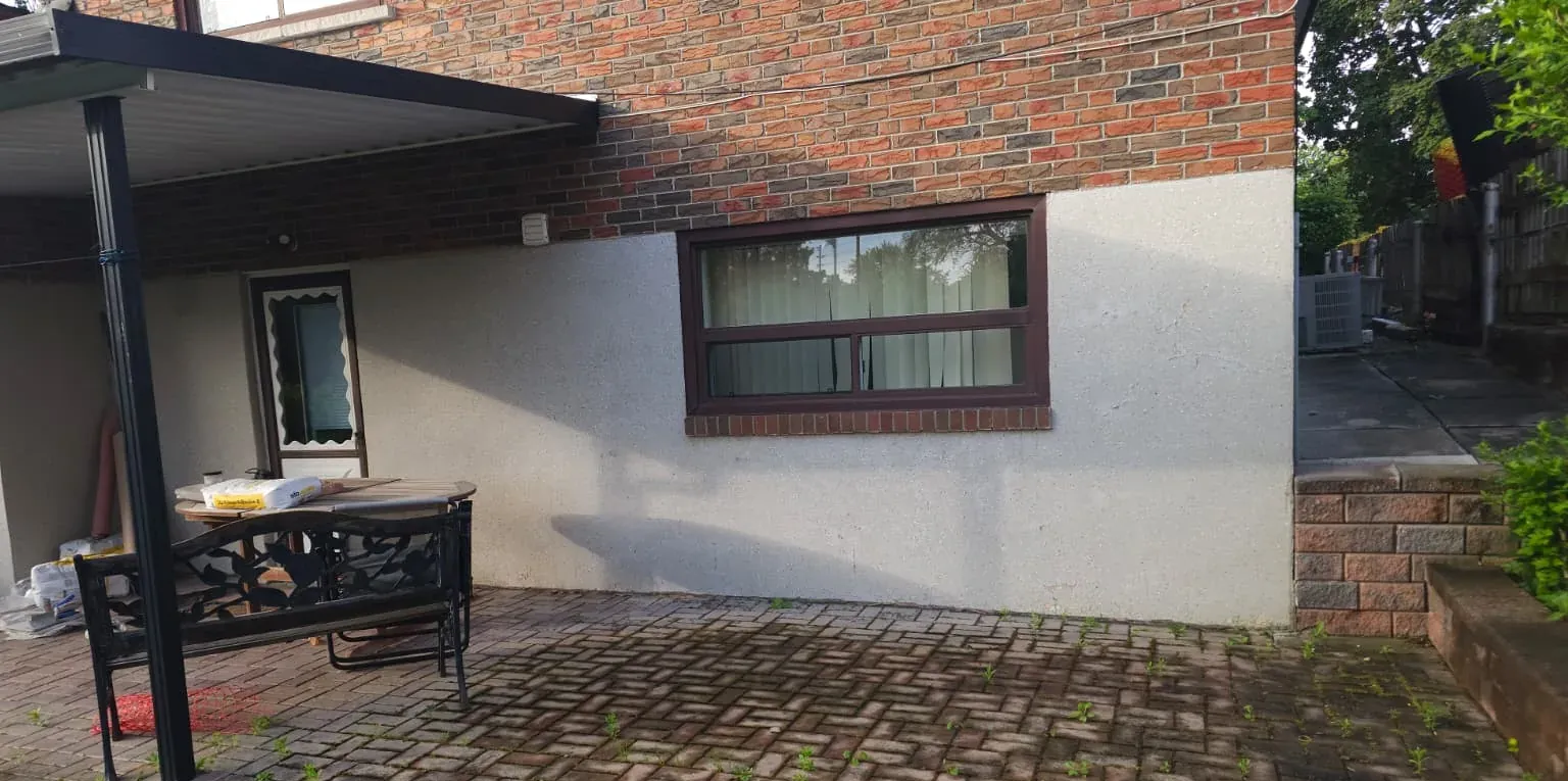 A shaded stucco wall and patio behind a house in Scarborough before FAIME refinished it, with growth in the damp paver joints