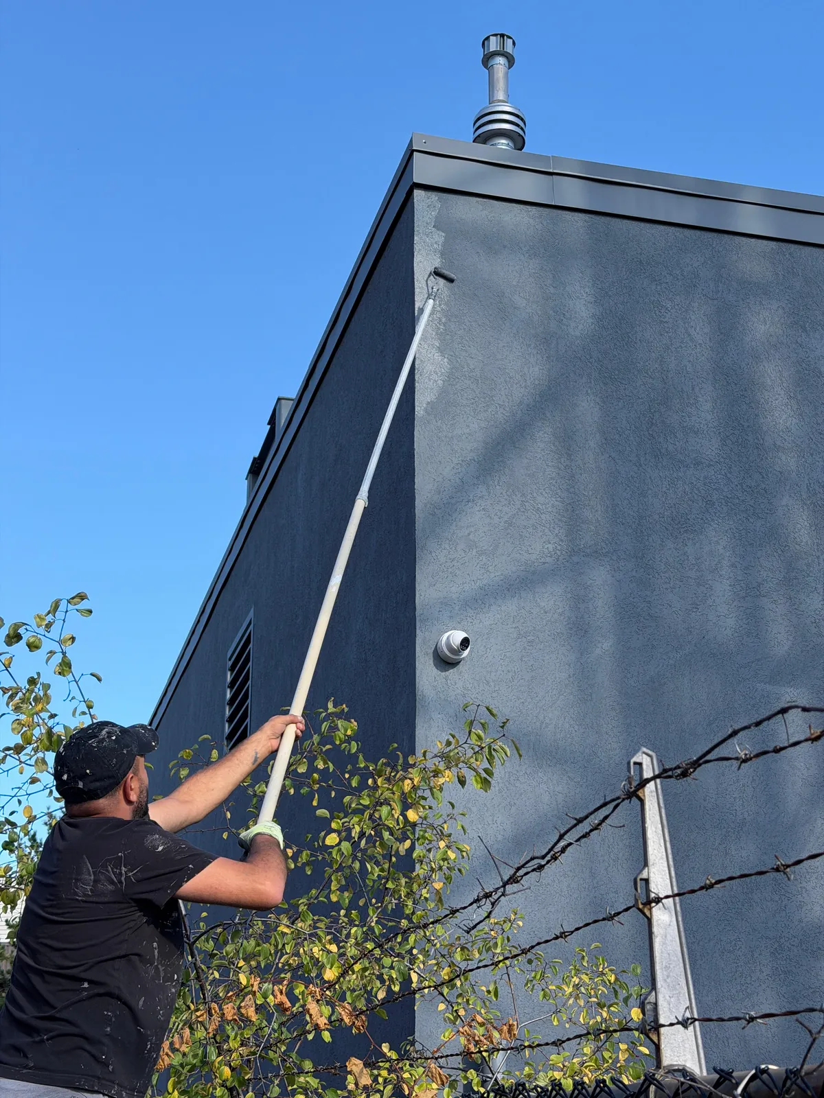 A FAIME painter rolling the upper wall from the ground on an extension pole, beside a perimeter fence