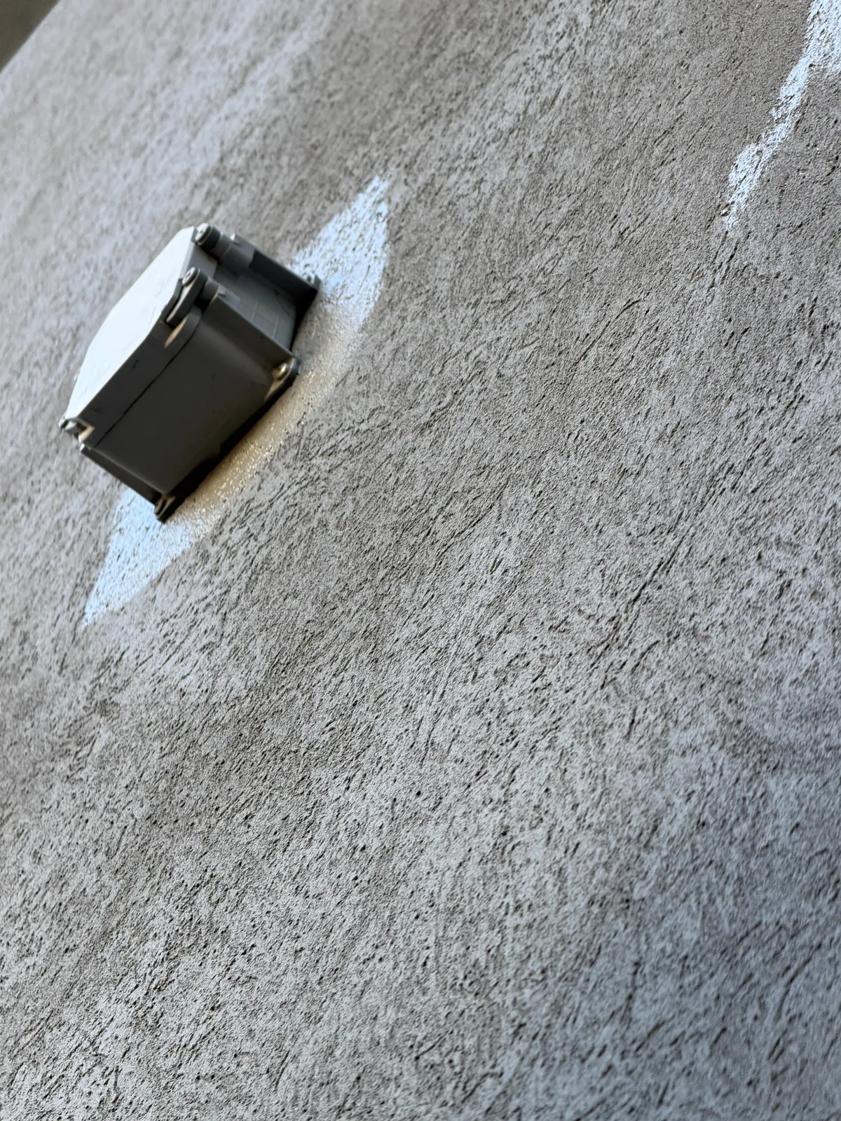 A wall-mounted electrical box with the grey being cut in around it on the textured stucco