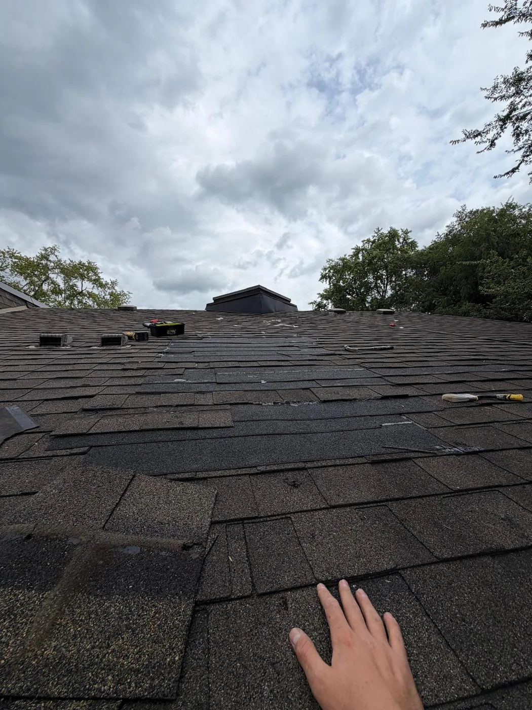 A hand resting on lifted and torn asphalt shingles on the roof slope above the leak.